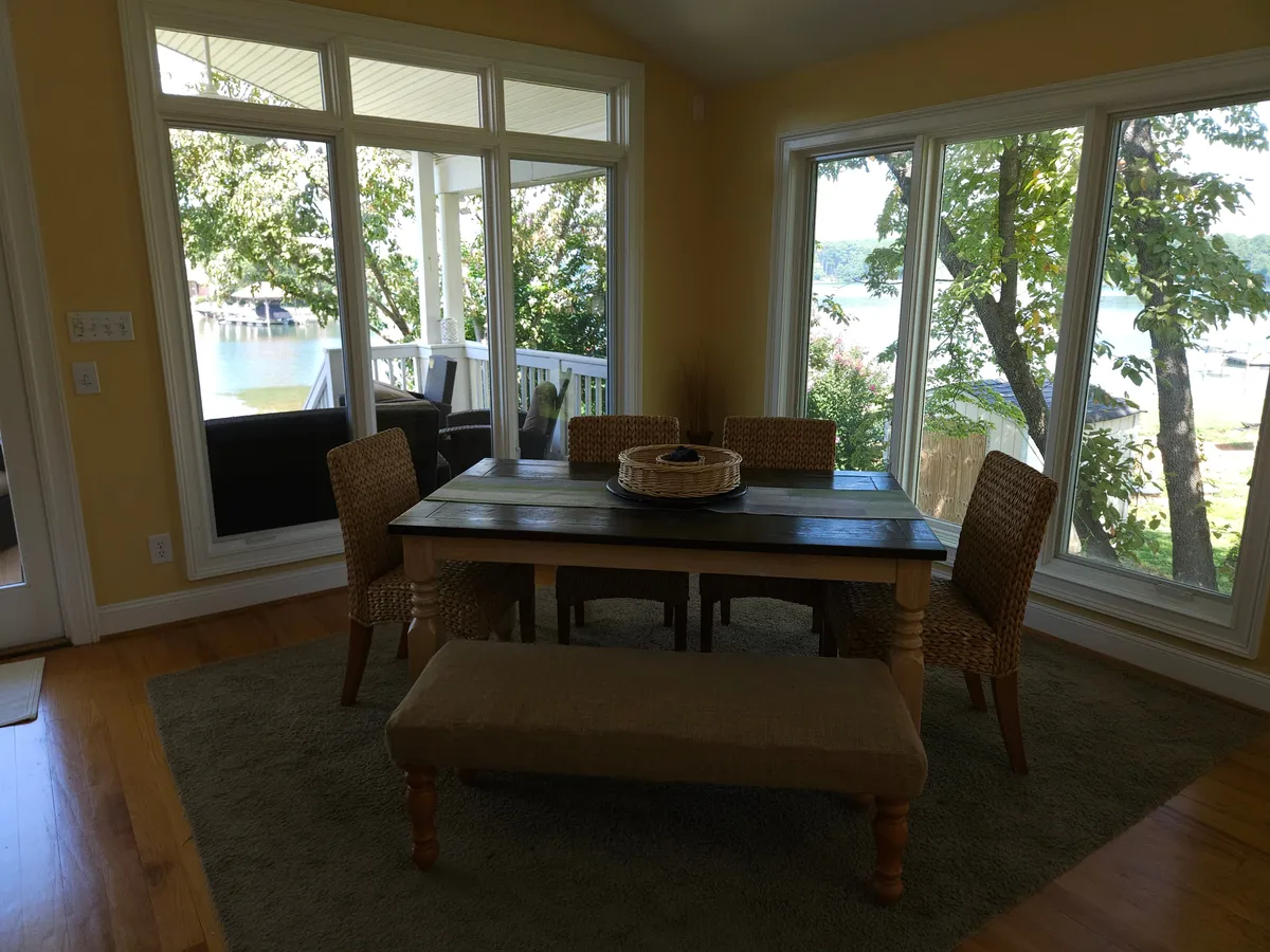 A cozy dining area with woven chairs is set next to large windows that offer panoramic views of the water. The space is styled for informal family meals or entertaining, with a relaxed and comfortable ambiance.