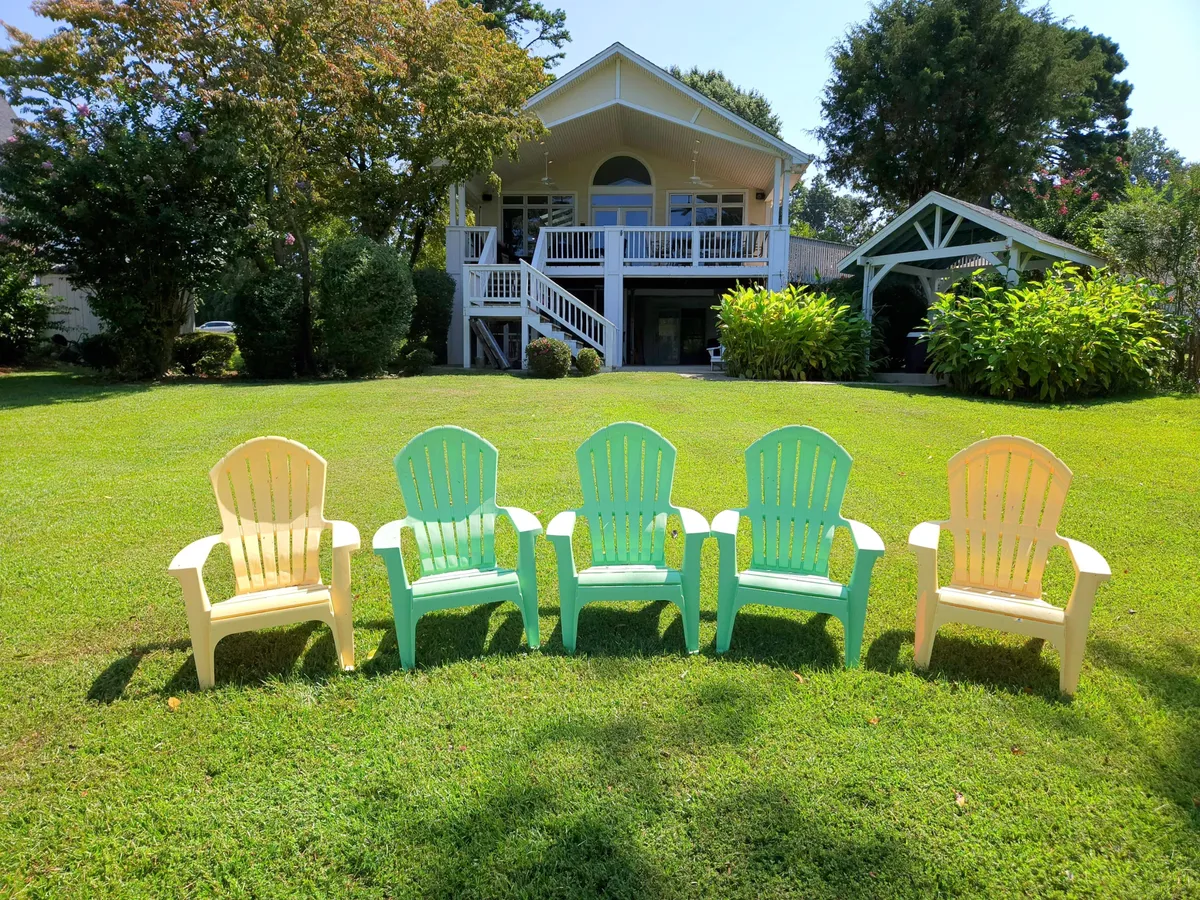 This image shows a collection of colorful Adirondack chairs arranged on a lush green lawn, with the house visible in the background. The chairs are oriented to take in the natural surroundings, suggesting a relaxing outdoor atmosphere adjacent to the lake.