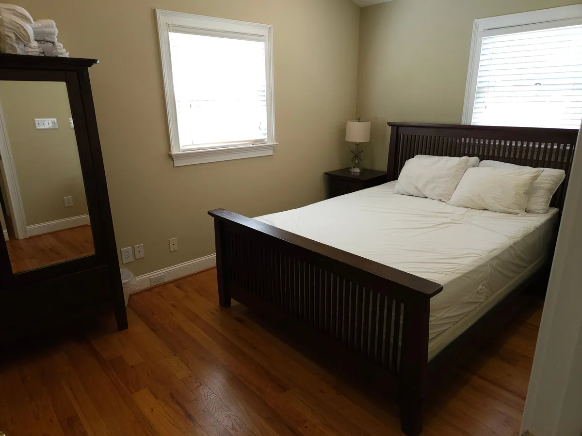 A serene bedroom featuring a cozy bed with soft linens, a nightstand with a reading lamp, and a mirror reflecting gentle sunlight. The simple, inviting decor creates a peaceful atmosphere ideal for restful nights.