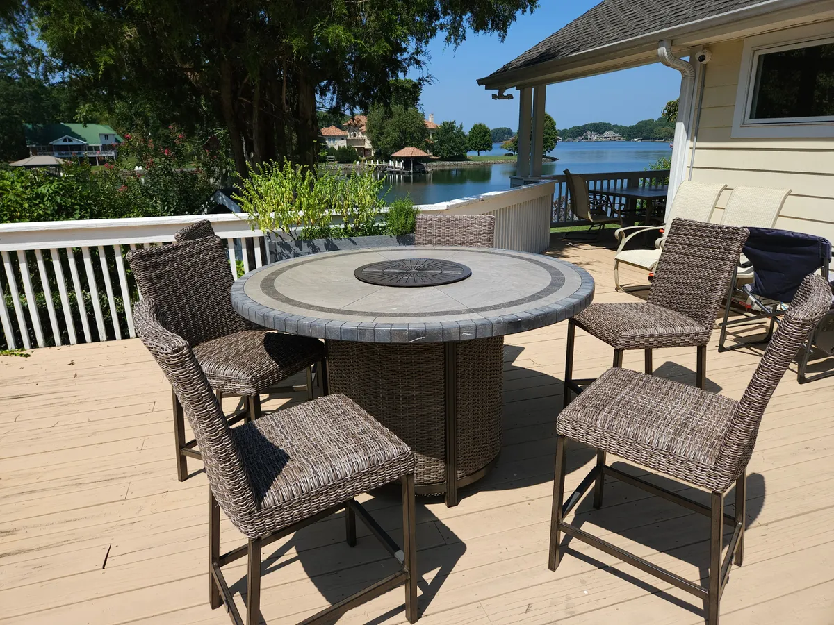 A comfortable outdoor dining area with a table and wicker chairs overlooks the lake, offering sweeping water views. The setting radiates comfort and is ideal for enjoying meals, drinks, or simply soaking up the tranquil scenery.