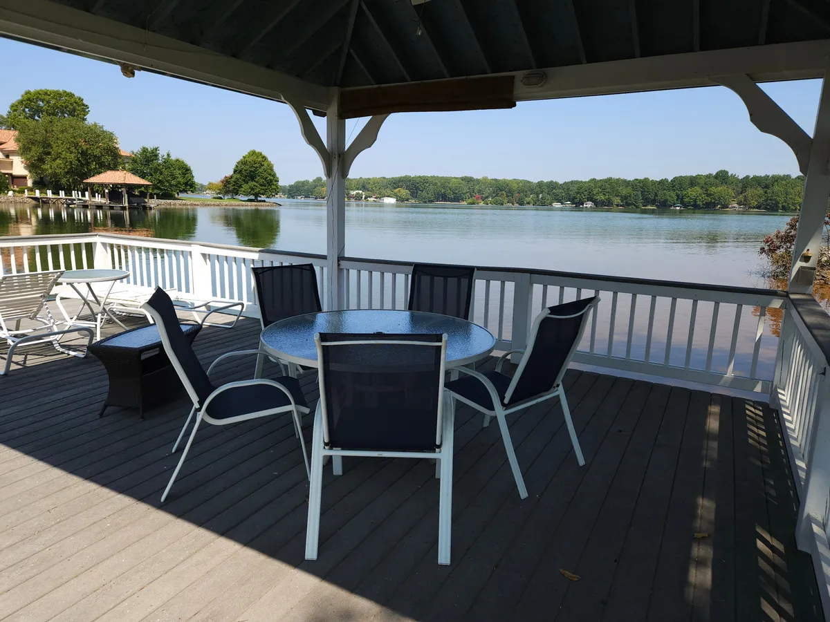 This exterior image features a round dining table and chairs set on a deck surrounded by trees, with a beautiful view of Lake Norman. The inviting space is ideal for outdoor dining or relaxing, with the shimmering water visible beyond the railing, capturing a tranquil lakefront atmosphere.