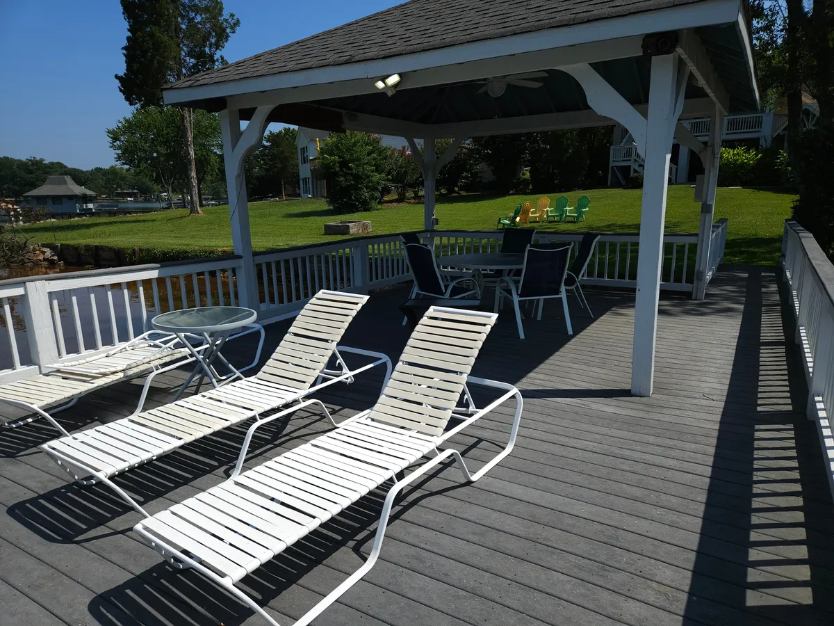 A spacious outdoor deck furnished with lounge chairs, a dining table, and colorful chairs in the background. The setup invites both sunbathing and outdoor meals, set against the backdrop of the water and treed shoreline.