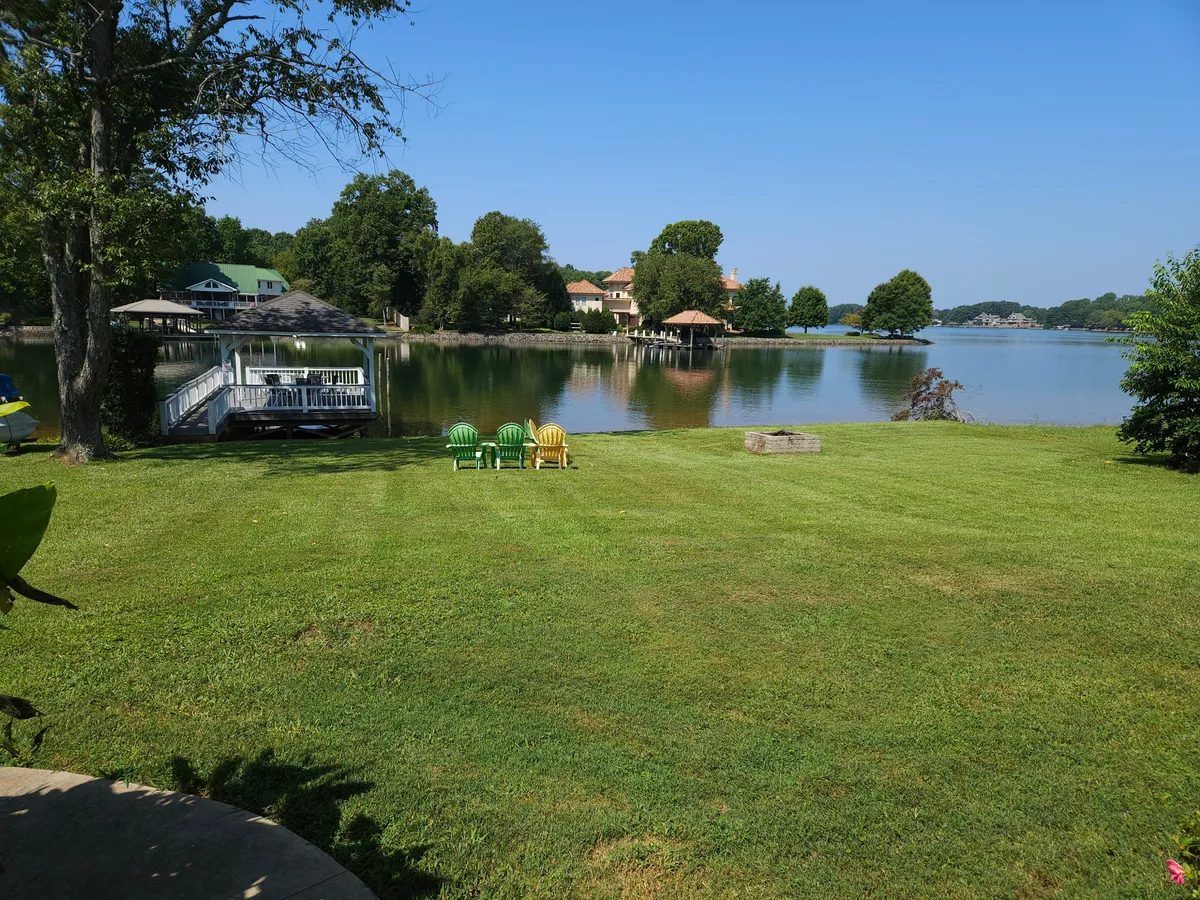 An exterior shot of Lake Norman taken from the shore, showing a private dock with lawn chairs positioned for relaxation. The calm water, nearby houses, and lush trees add to the peaceful setting, inviting guests to lounge or fish by the lake.