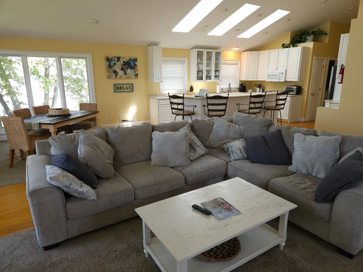 This image captures an open-concept living space that seamlessly combines the living room, dining area, and kitchen. Large windows along the back wall provide beautiful views of the lake and flood the room with natural light. The sectional sofa and dining table create inviting zones for family gatherings and entertaining guests.
