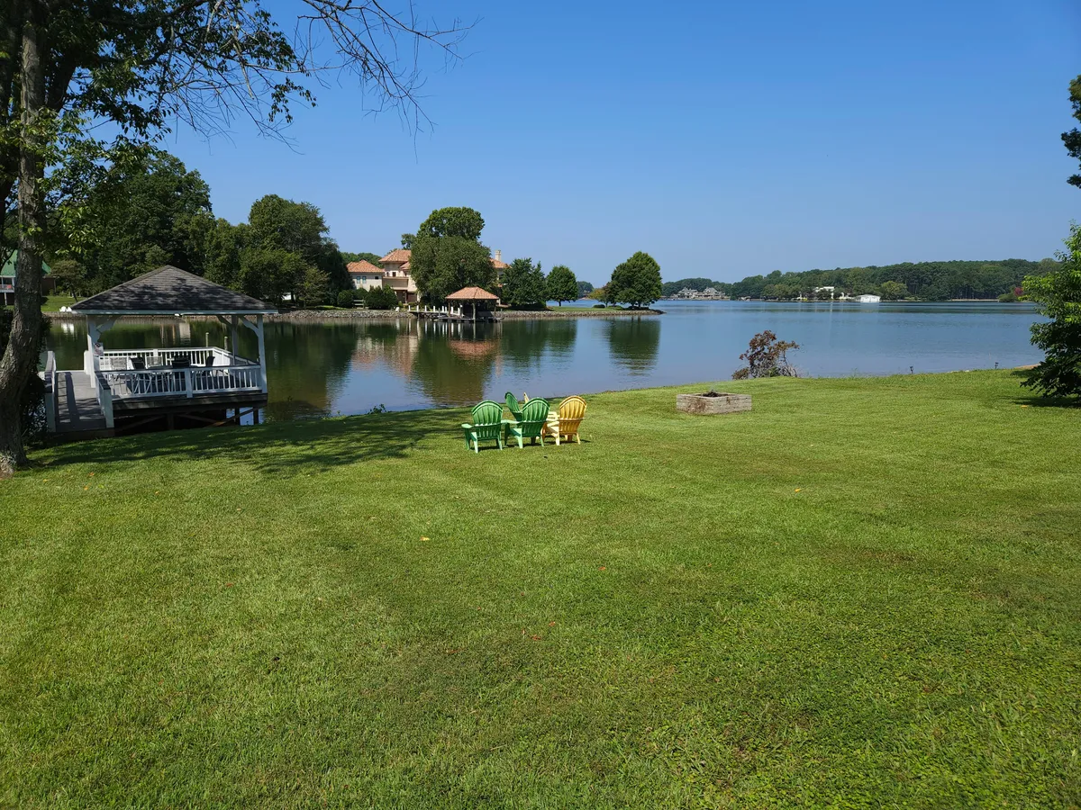 This image features colorful Adirondack chairs arranged around a gazebo near the calm surface of Lake Norman. The lakeside setting, with open lawn and water in the background, evokes a serene atmosphere perfect for evening gatherings and bonfires.