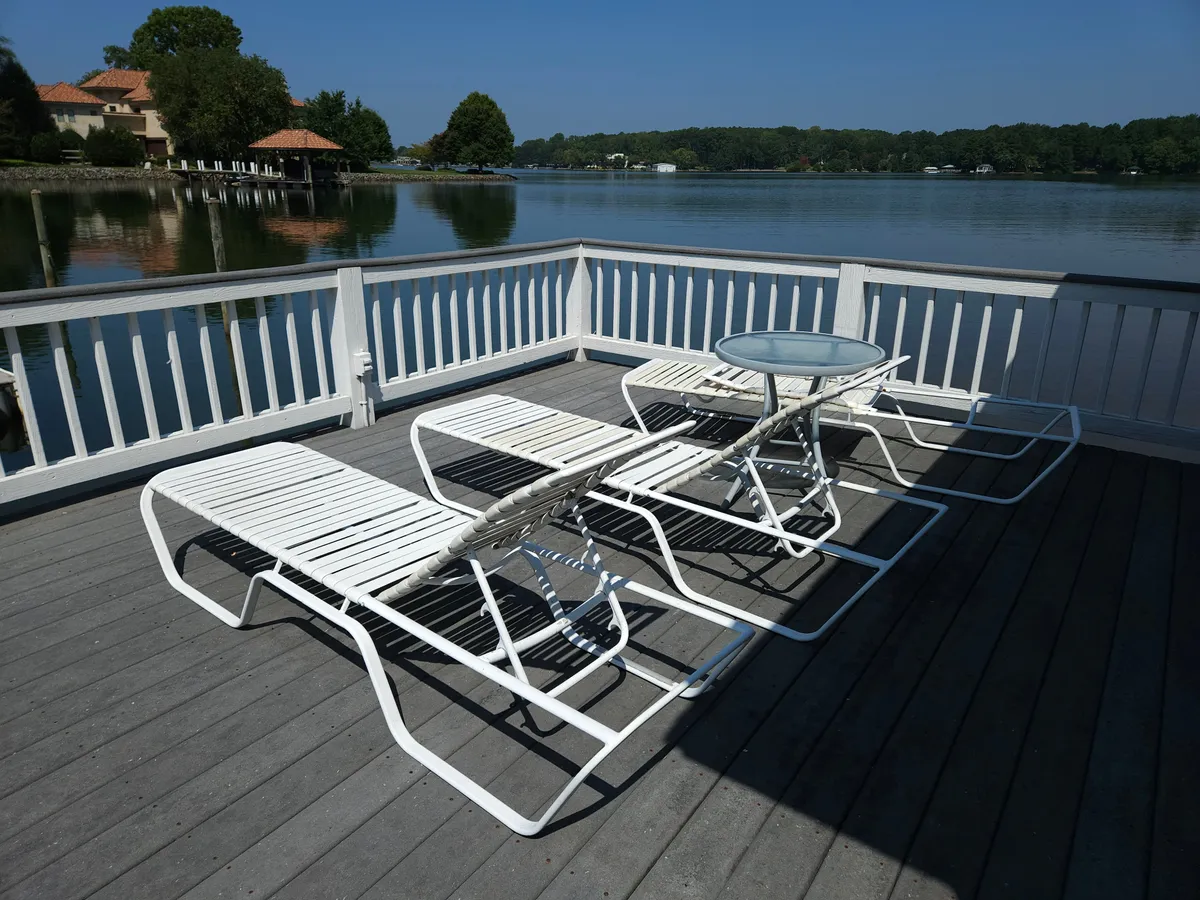 The image showcases a serene lakefront deck with inviting lounge chairs and a small table, perfectly positioned for guests to bask in the sun or unwind with a book. The scene opens up to the tranquil waters of Lake Norman, evoking relaxation and the promise of peaceful moments surrounded by nature.